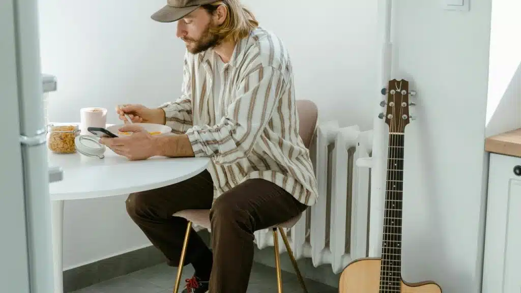 A man eating cereal at a table while looking at his phone with a guitar by the wall.