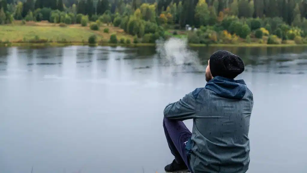 A man wearing a beanie sitting by a lake with trees in the background.