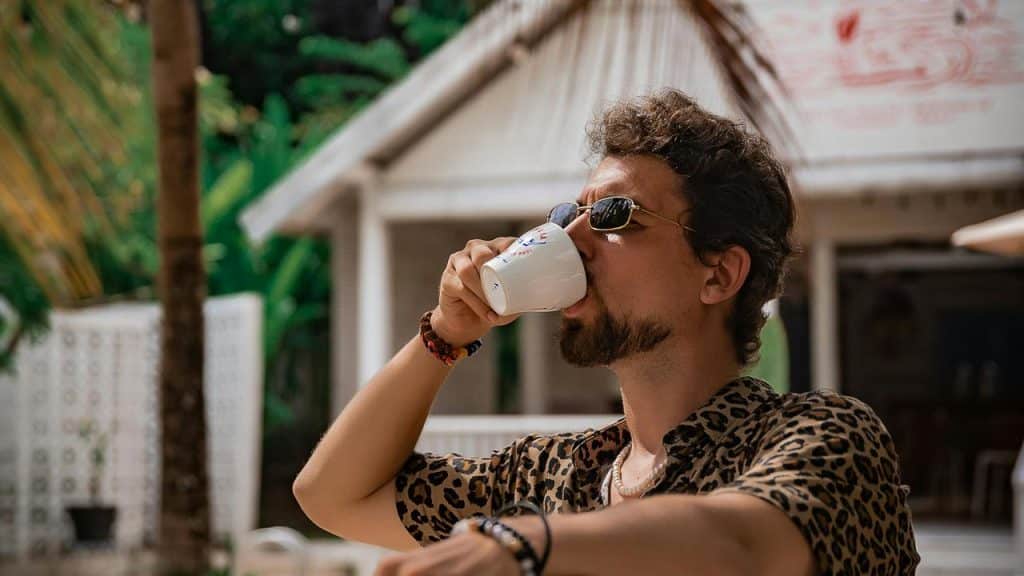 A man wearing sunglasses and a leopard-print shirt drinking from a white cup outdoors.