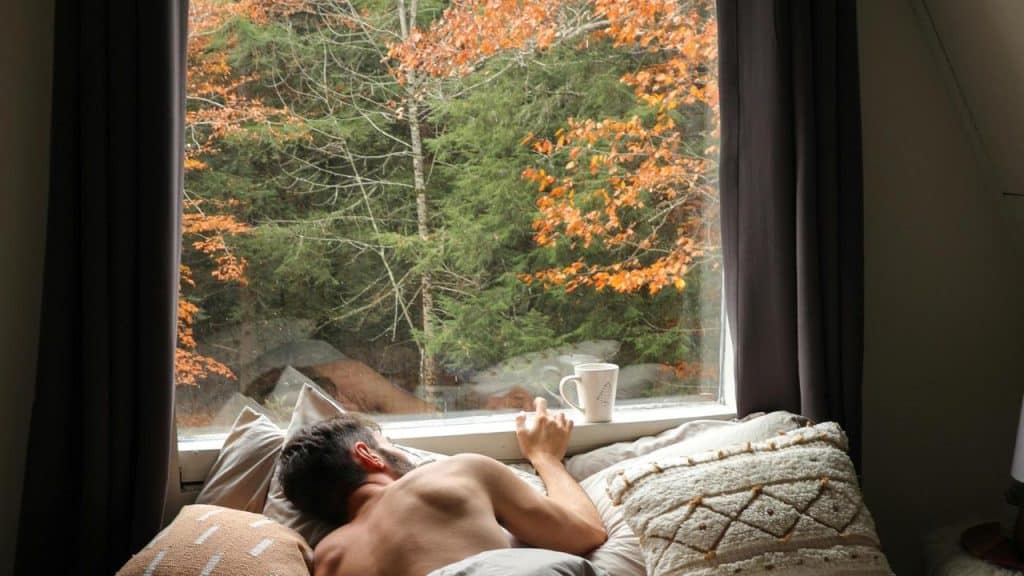 A man lying in bed by a window with a cup of coffee on the sill and autumn trees outside.