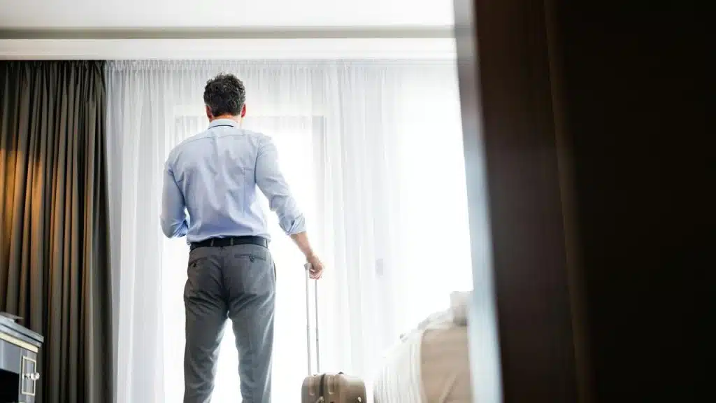 A man standing by a window with a suitcase in a hotel room.