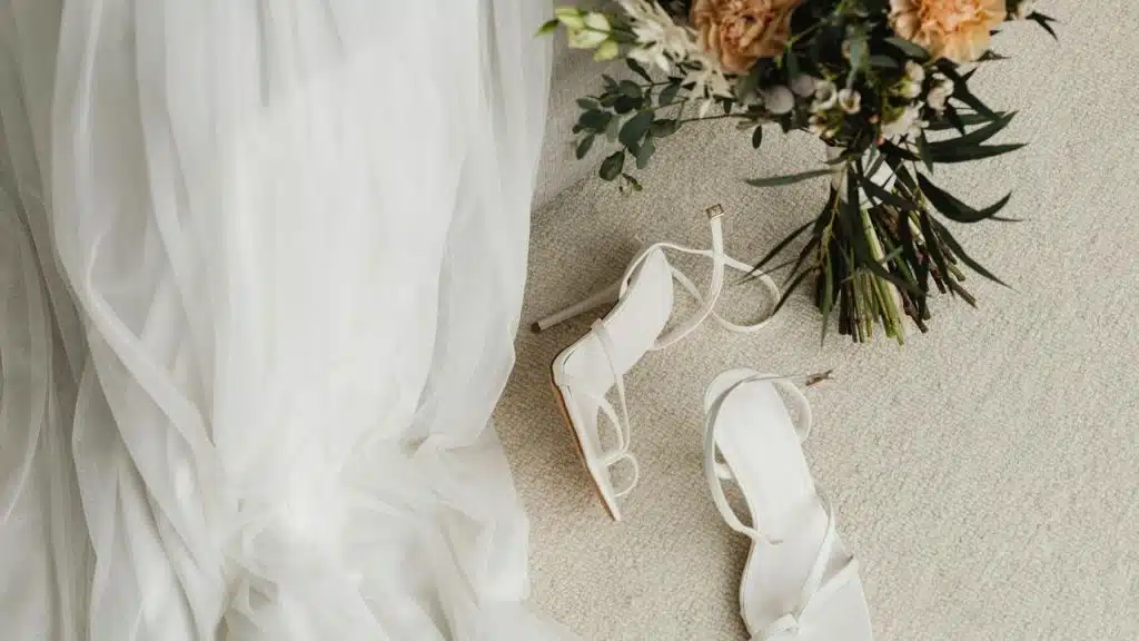A pair of white heels beside a wedding dress and bouquet.