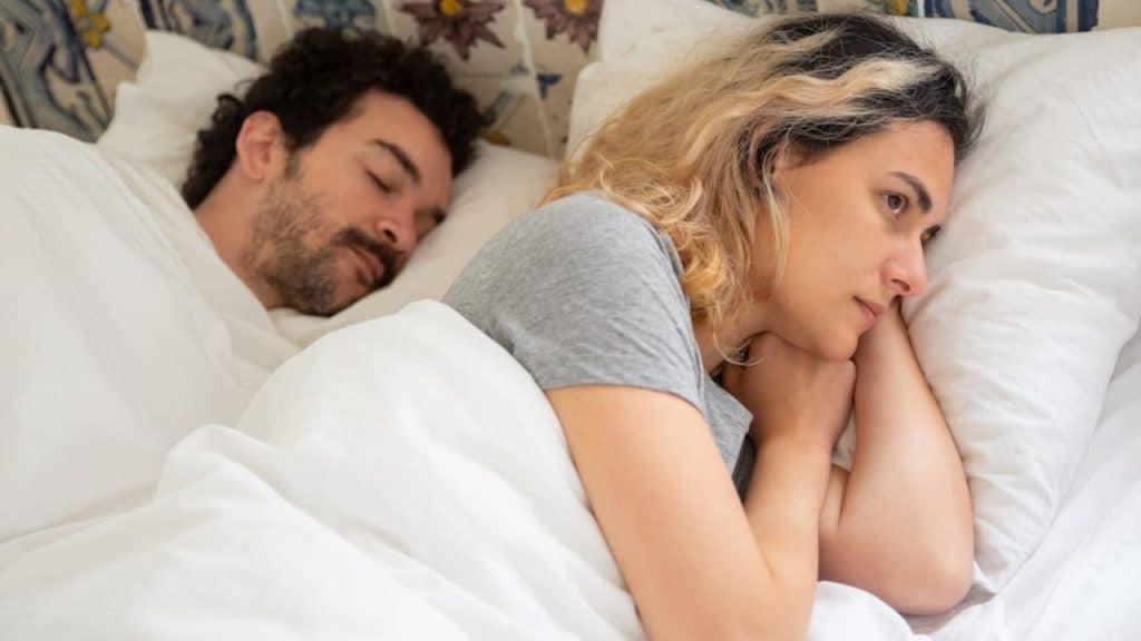 A blonde woman is awake and worried in bed next to a sleeping man.