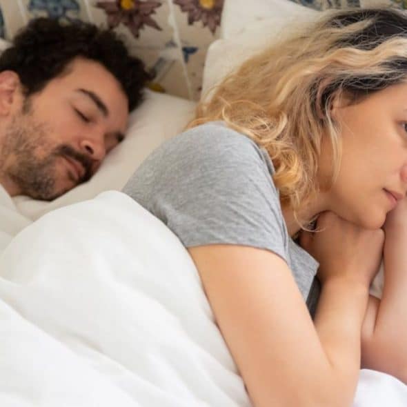 A blonde woman is awake and worried in bed next to a sleeping man.