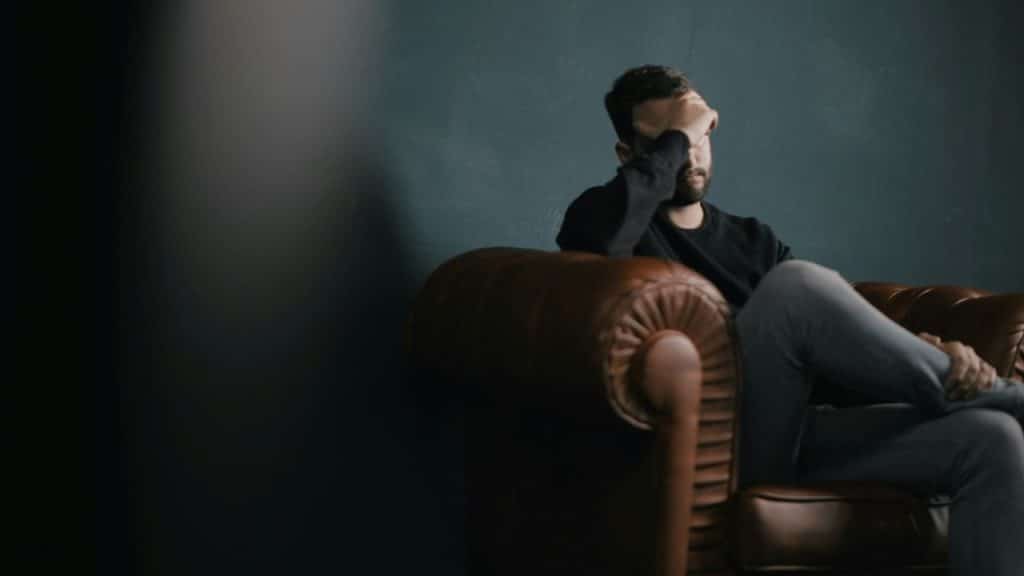 Man in black shirt sitting on leather couch, covering his eyes with his hand.