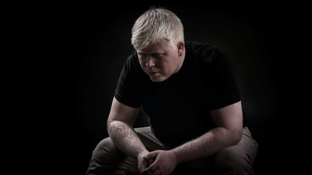 Man with white hair, wearing a black shirt, sitting in dark lighting with hands clasped.