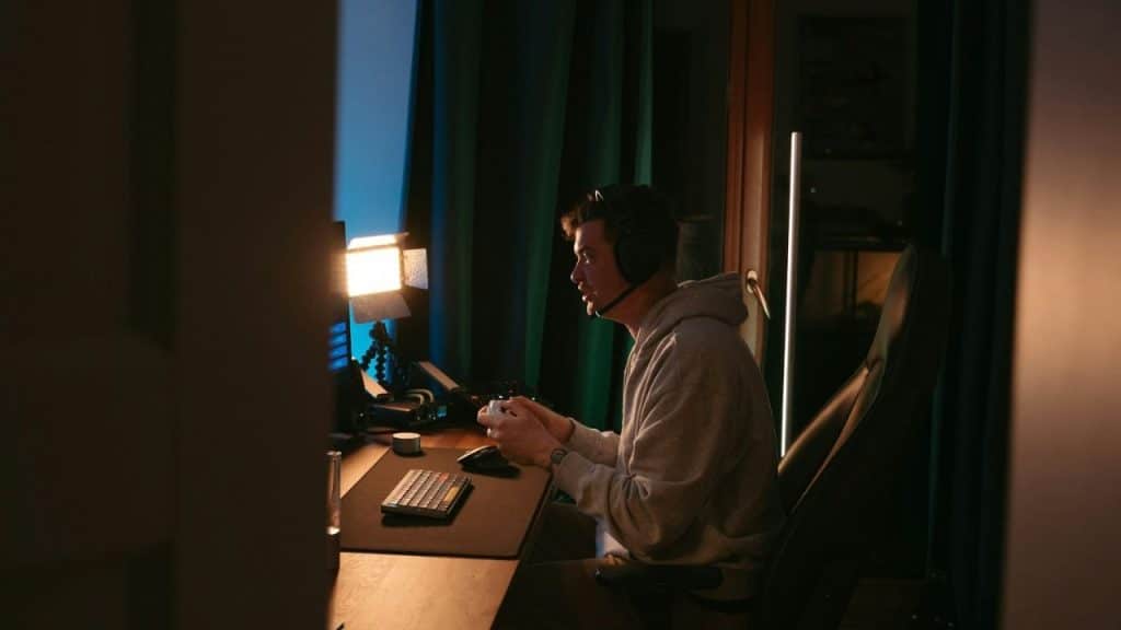 A man in a hoodie plays a video game on a computer in a dim room.