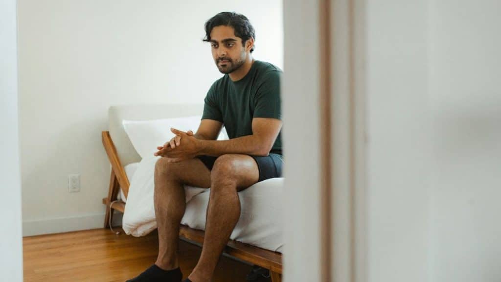 Man in dark shirt and shorts sitting on a bed with hands clasped in front.