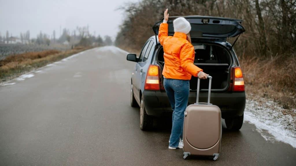 A woman stands by a car trunk on a gray roadside holding a suitcase.