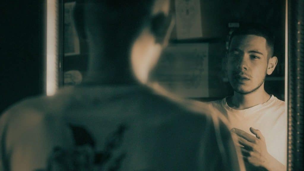 Man in white shirt looking at his reflection in a mirror, pointing at his chest.