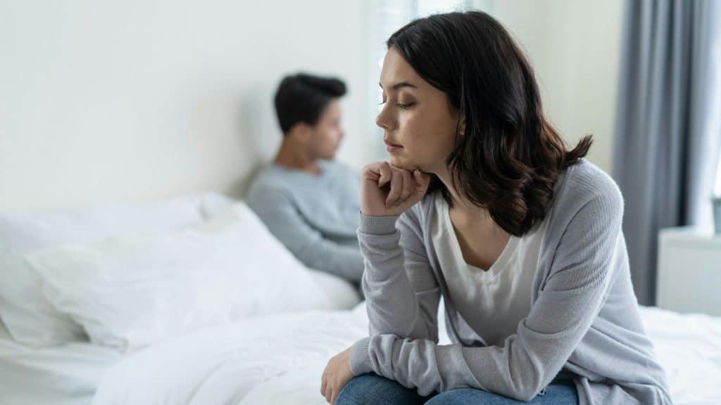 A young woman with her eyes closed sits on a white bed, her chin resting on her hand, while a man sits facing away in the background.