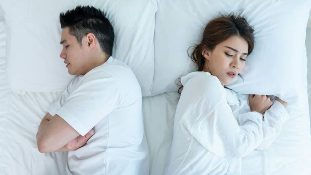 A man and woman lie in bed, back-to-back, with their arms crossed, looking distressed.