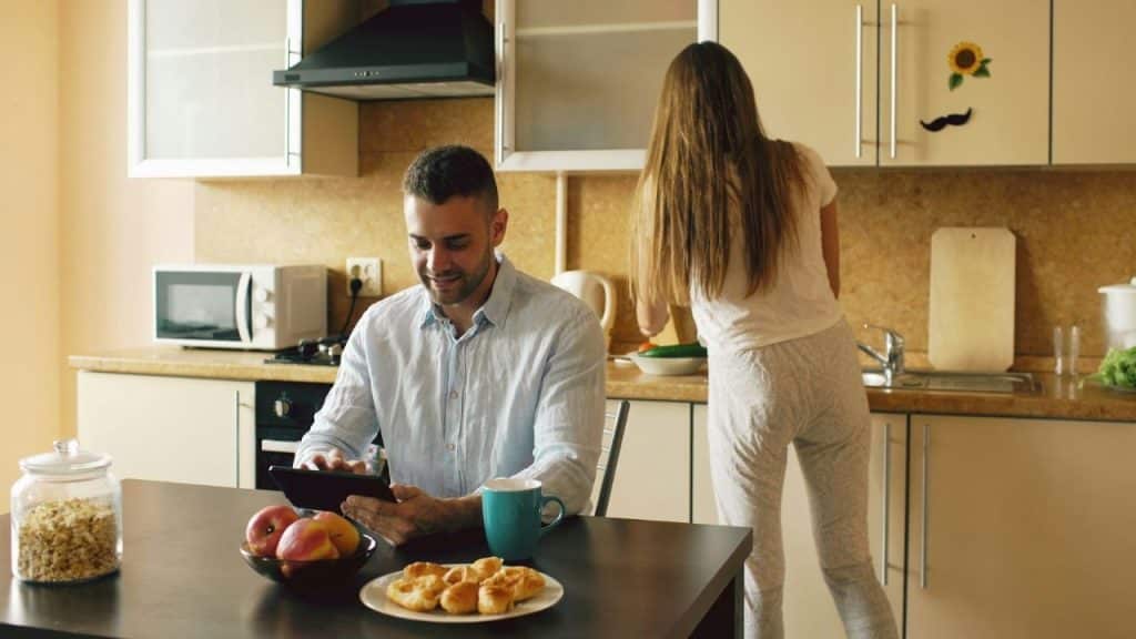 A man uses a tablet at a kitchen table while a woman stands with her back to him washing dishes.