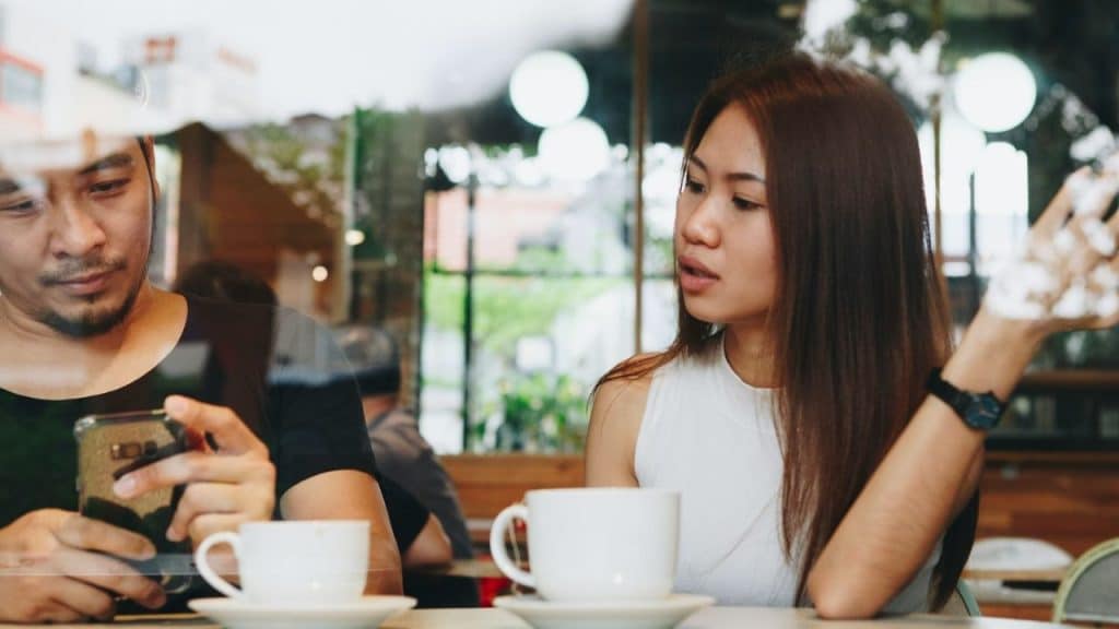 A man looks at his phone while a woman gestures and speaks across two coffee cups.