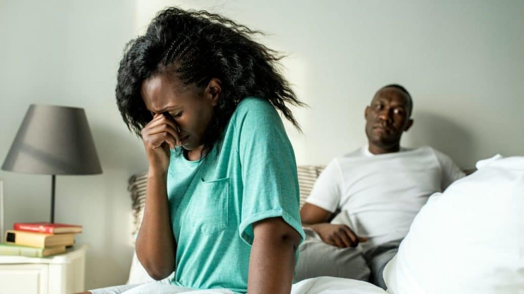 A Black woman sits on a bed with her hand over her face while a man sits behind her.