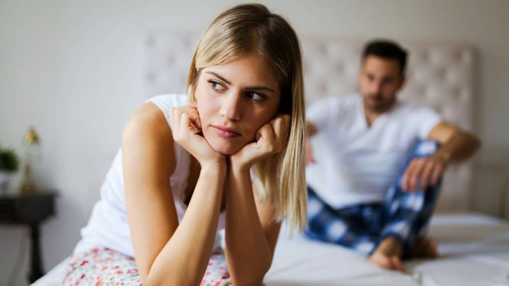 Woman sitting on a bed looking upset with a blurred man in the background.
