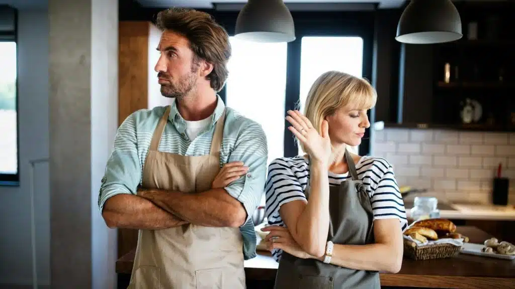 Man and woman in aprons standing back-to-back in a kitchen, looking frustrated.