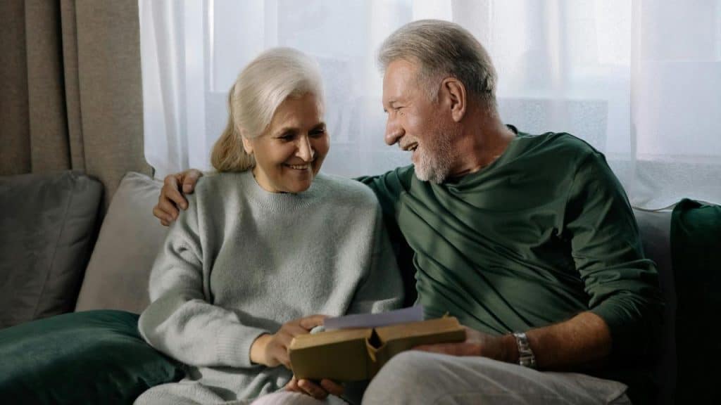 An older couple sitting together on a couch and smiling while holding a book.