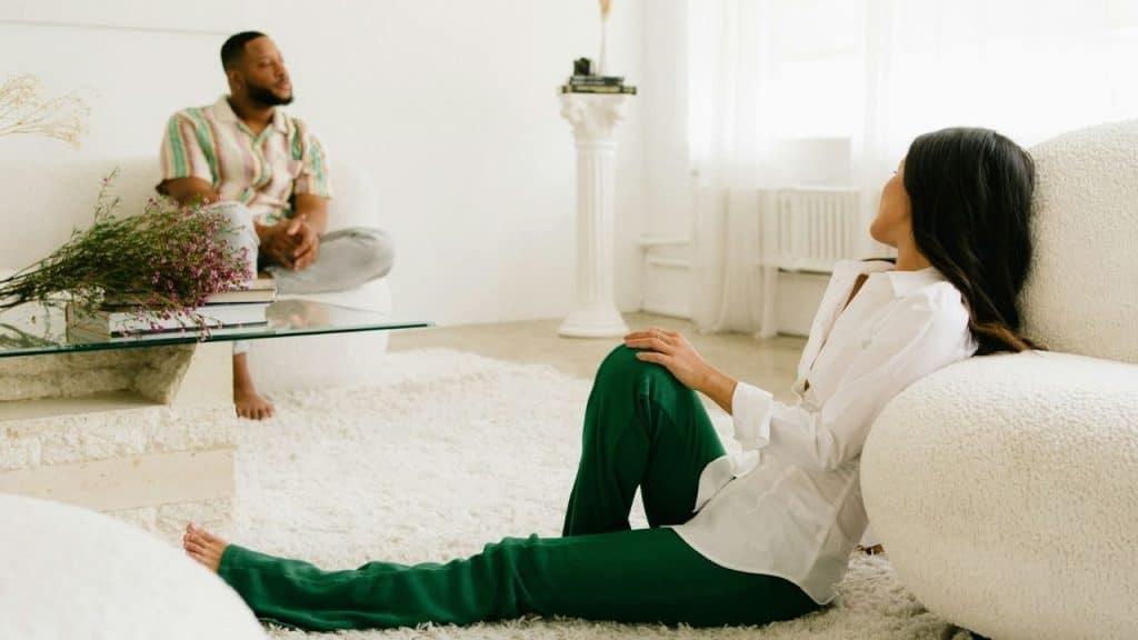 A woman in green pants sits on a white shaggy rug facing a man sitting on a couch.