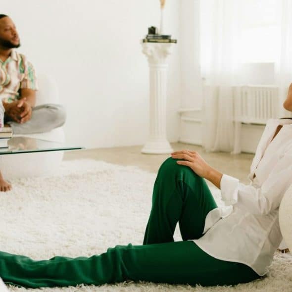 A woman in green pants sits on a white shaggy rug facing a man sitting on a couch.