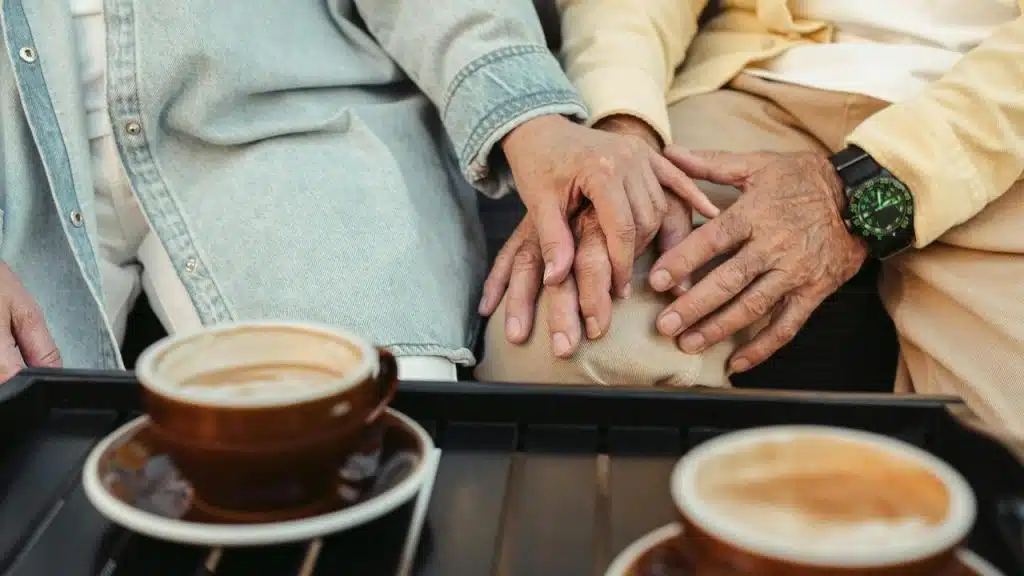 An older couple’s hands resting gently together beside coffee cups.