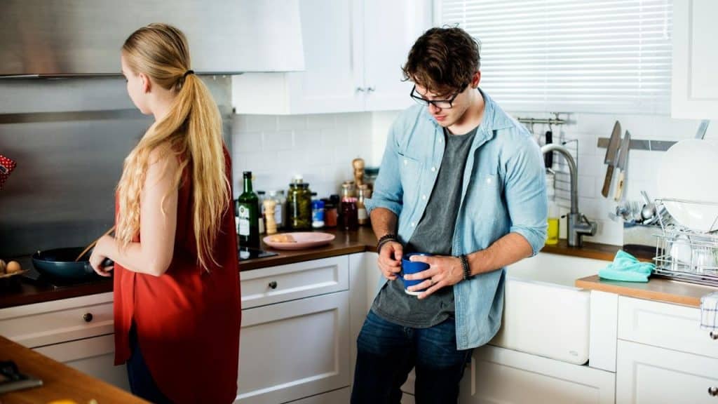 A man is looking down, holding a mug, while a woman cooks at a stove in a white kitchen.