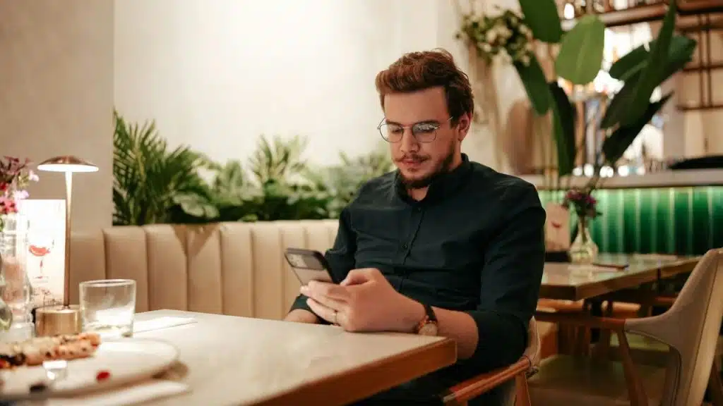 Man with glasses sitting at a restaurant table, looking down at his phone.