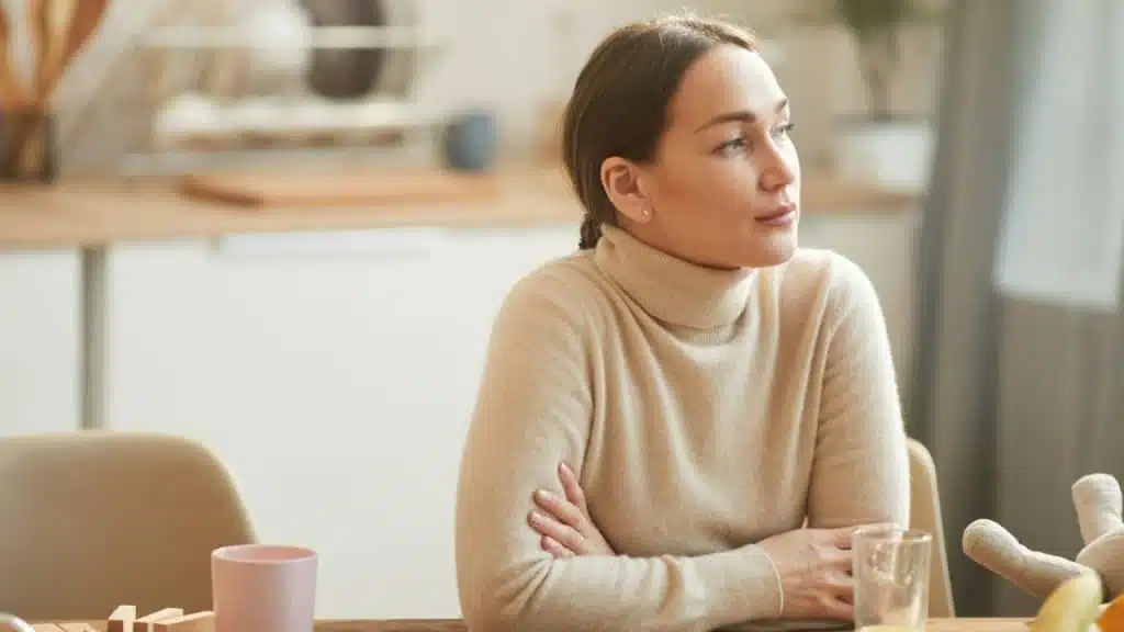 Woman sitting at a kitchen table with arms crossed, looking away from the camera.