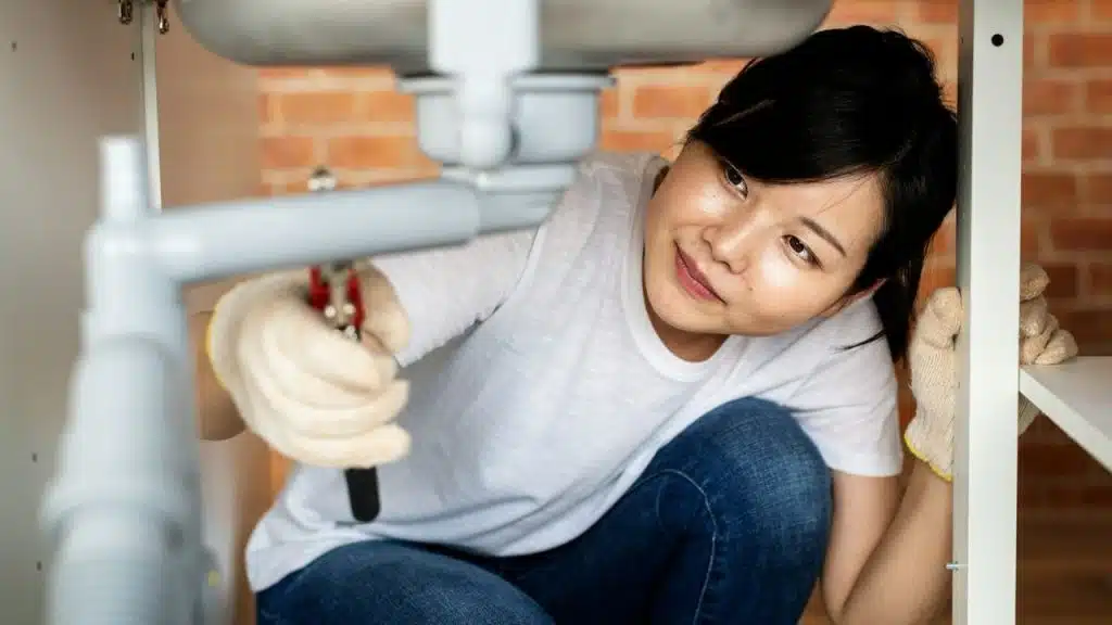 Asian woman in a white shirt and gloves fixing a pipe under a sink.