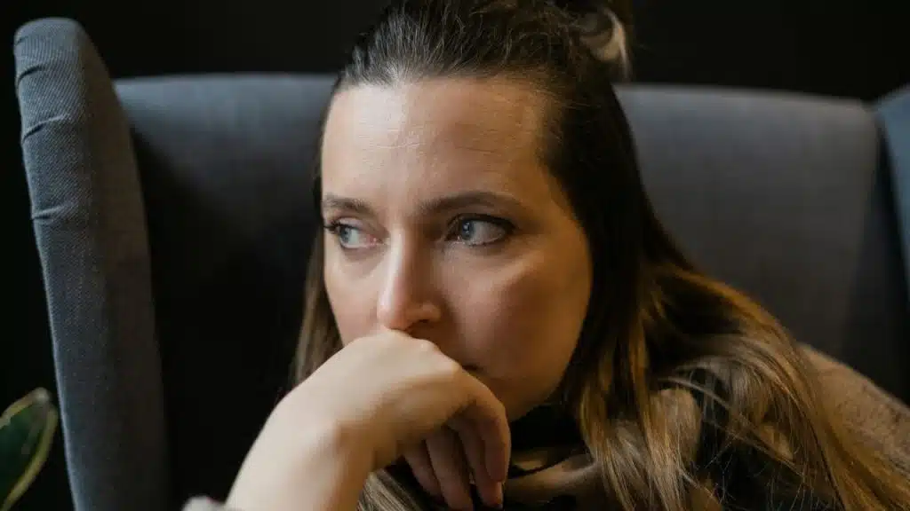 Woman in a gray armchair looking away, with her hand near her mouth.