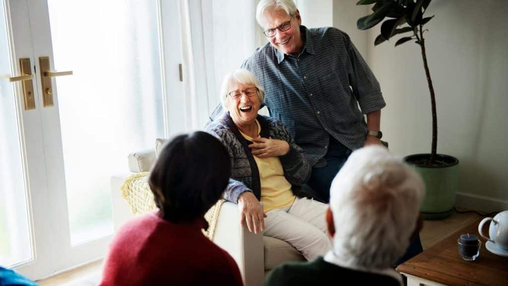 An older couple laughing and chatting with friends in a cozy living room.