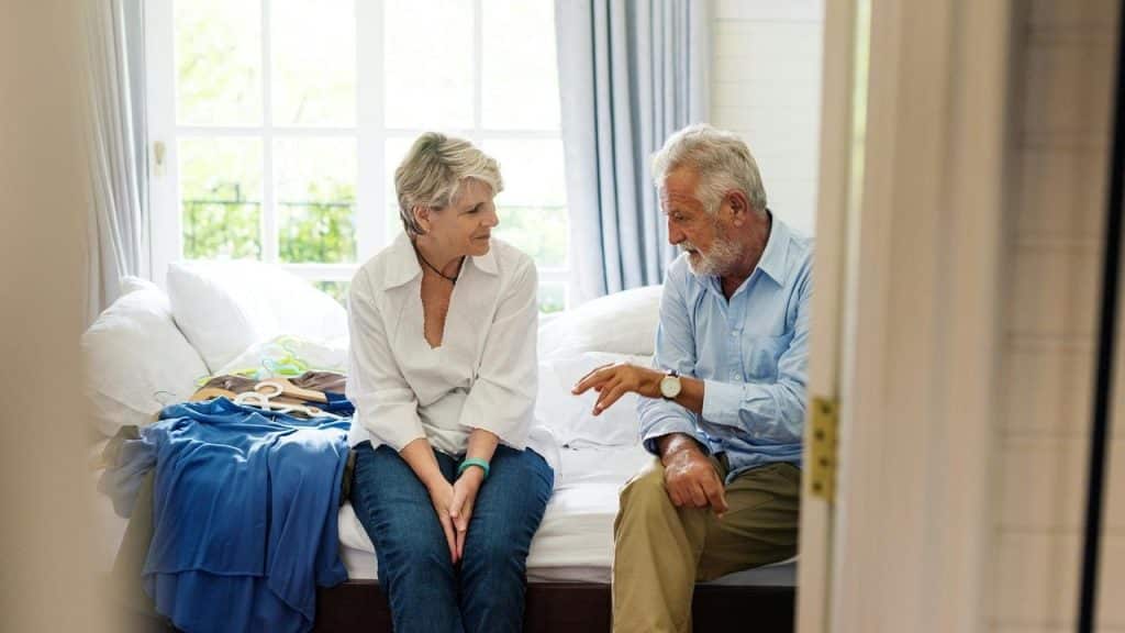 An older couple sitting on a bed and talking together.