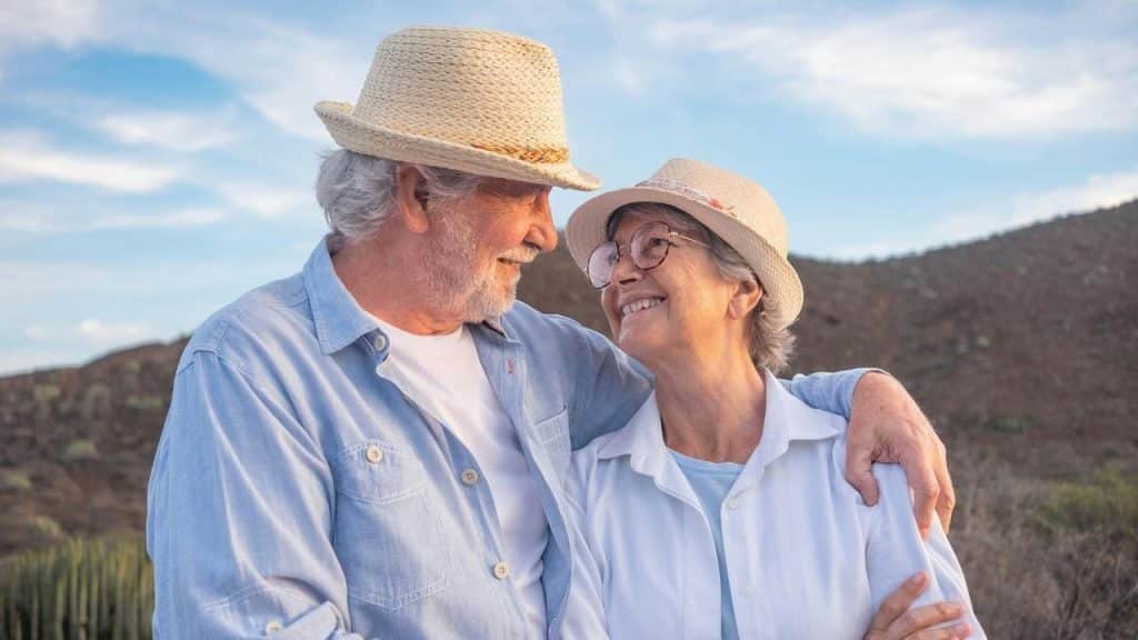 An older couple wearing hats and smiling at each other outdoors.