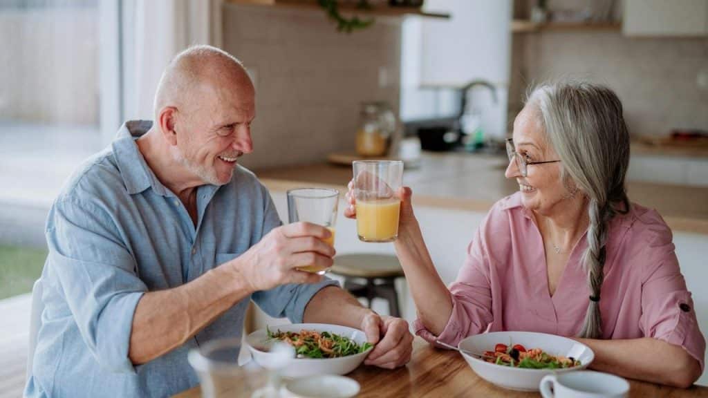 An older couple smiling and toasting with juice while eating a meal together.