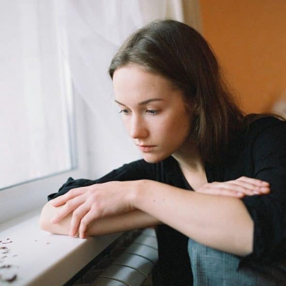 Woman with long brown hair looks down sadly, resting her arms on a windowsill.