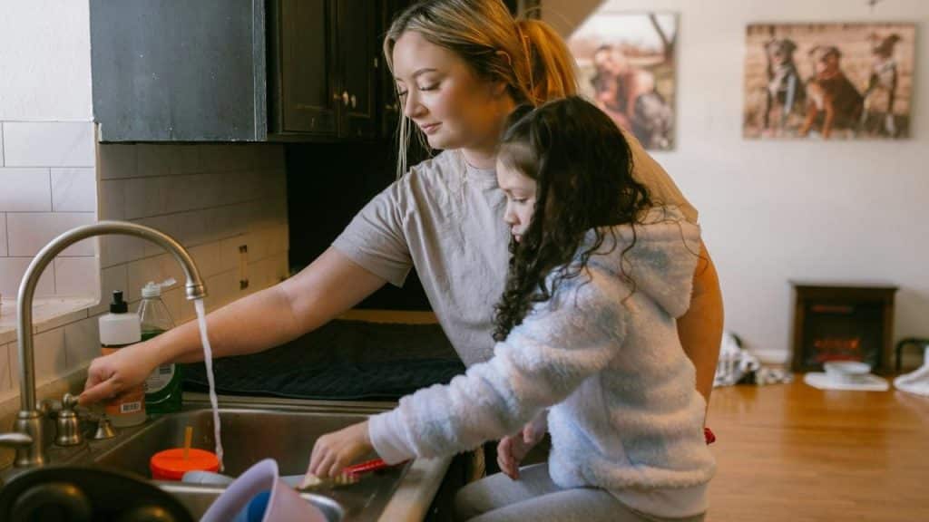 A woman helping a young girl wash dishes at a kitchen sink.