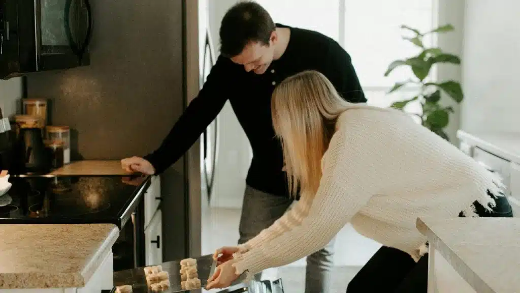 A couple placing a tray of cookie dough into an oven in their kitchen.