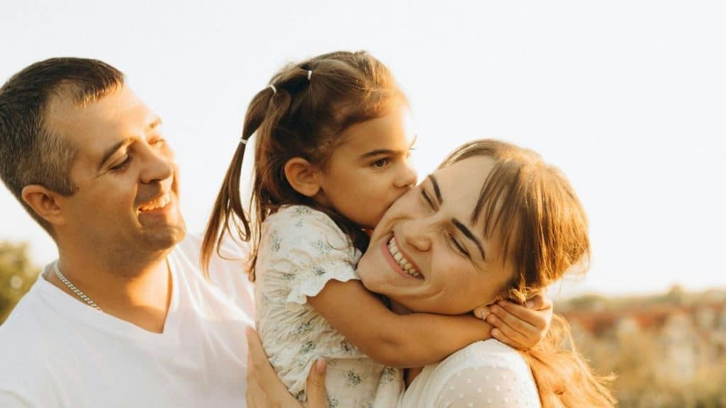 A young girl hugging and kissing a smiling woman while a man looks on happily.