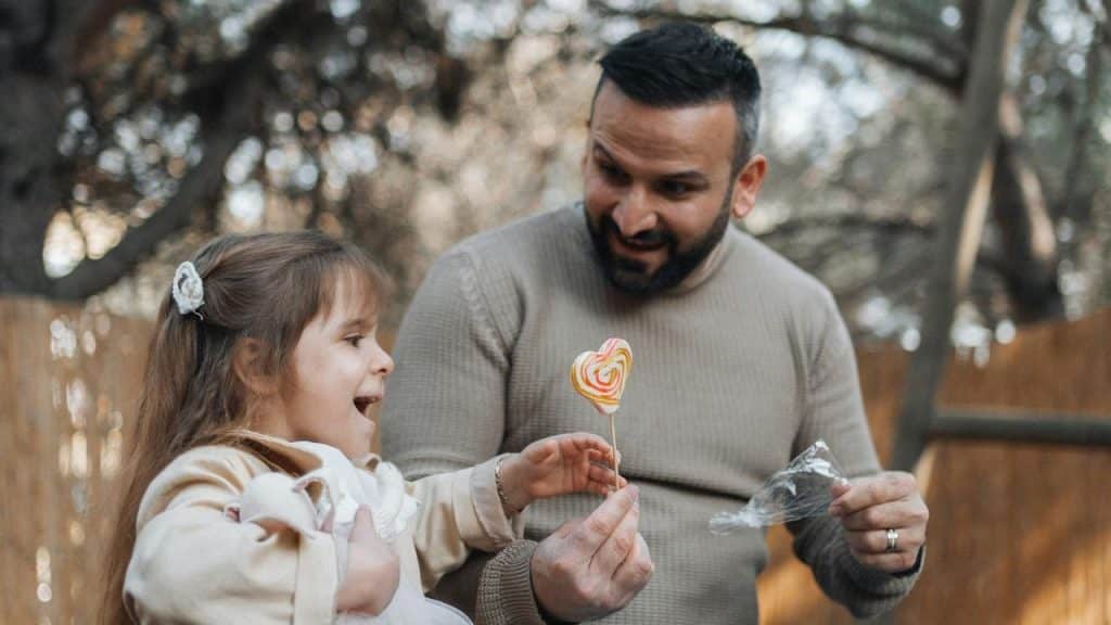 A man giving a young girl a heart-shaped lollipop outdoors.