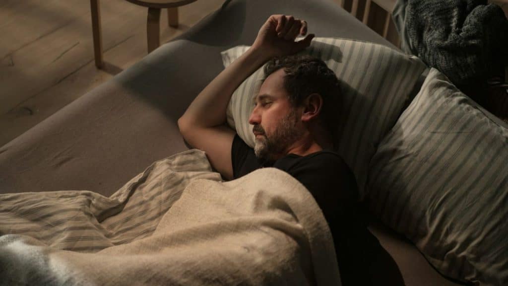 Middle-aged man lying in bed, asleep or resting, with his arm over his head.