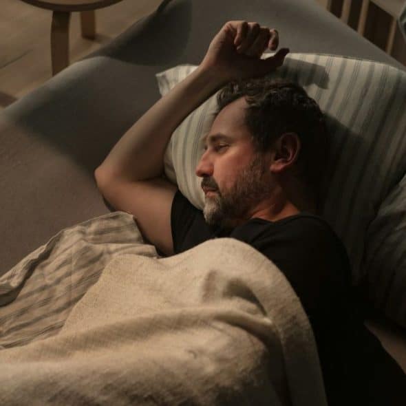 Middle-aged man lying in bed, asleep or resting, with his arm over his head.