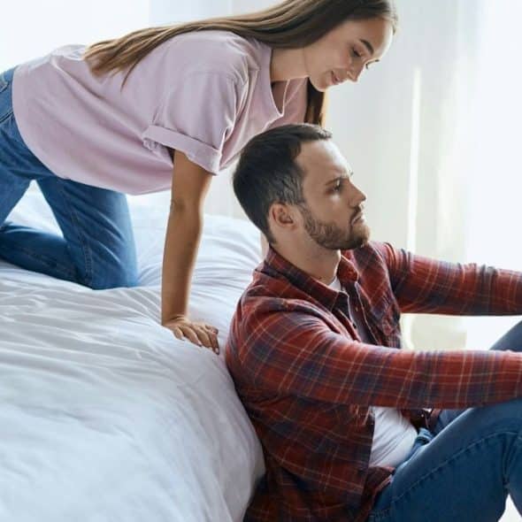 A man sitting on the floor looking away from a woman kneeling on the bed.