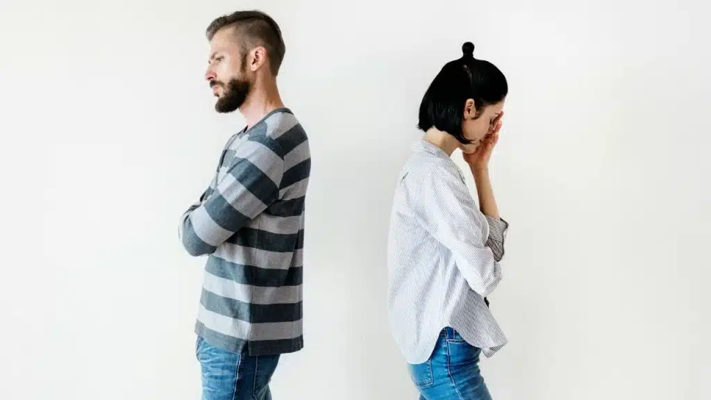A man and a woman stand back-to-back with crossed arms against a plain white wall.