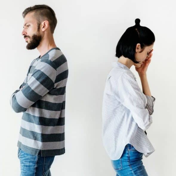 A man and a woman stand back-to-back with crossed arms against a plain white wall.
