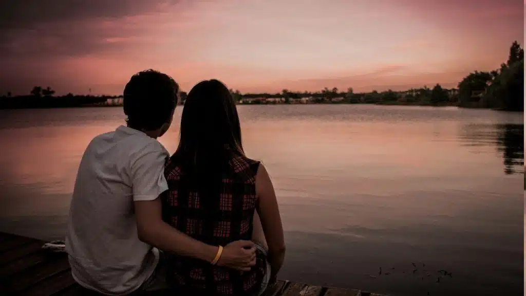 A couple watching the sunset while sitting by a lake’s shore.