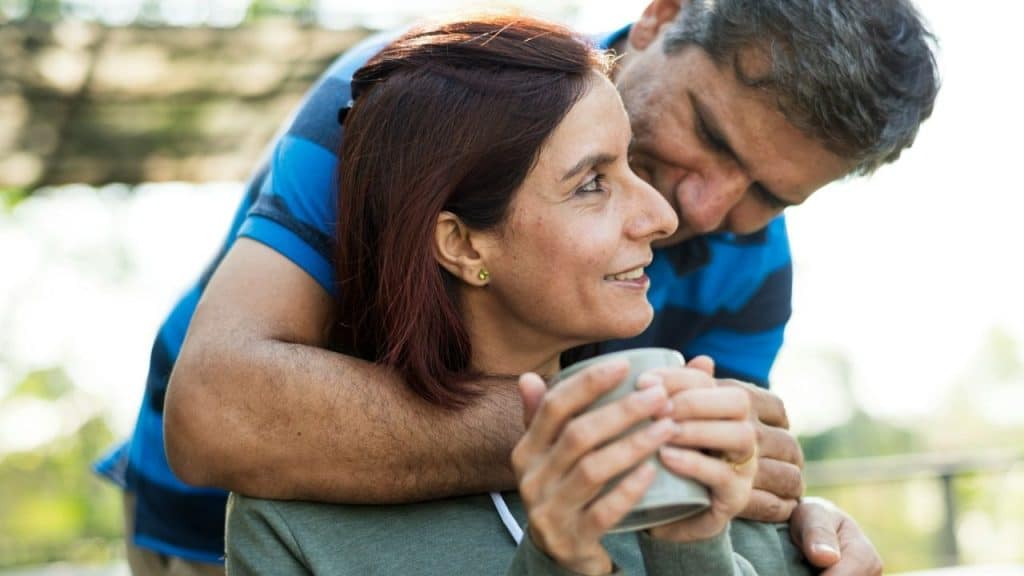 A middle-aged man is embracing a woman from behind who is holding a mug and smiling.