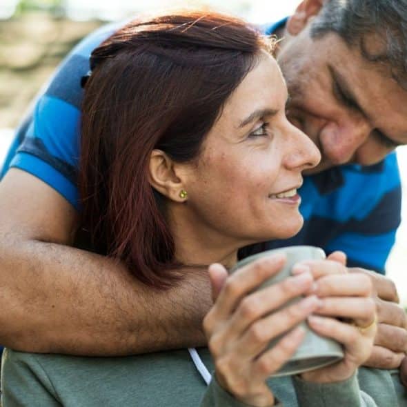 A middle-aged man is embracing a woman from behind who is holding a mug and smiling.
