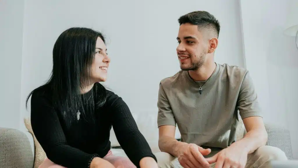 Smiling man and woman sitting together on a light-colored couch, looking at each other.