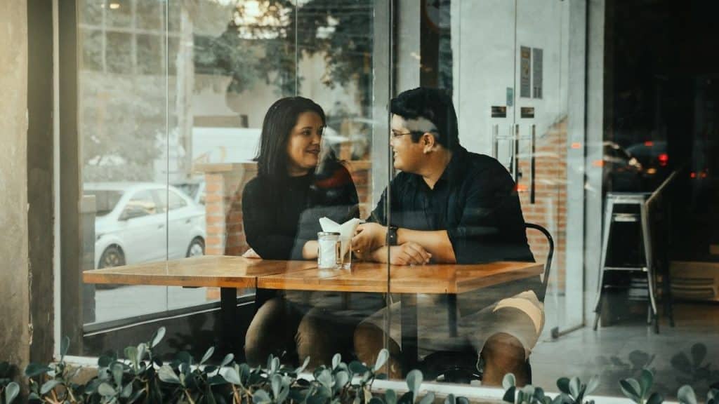 A man and woman are sitting at a cafe table, smiling and looking at each other.