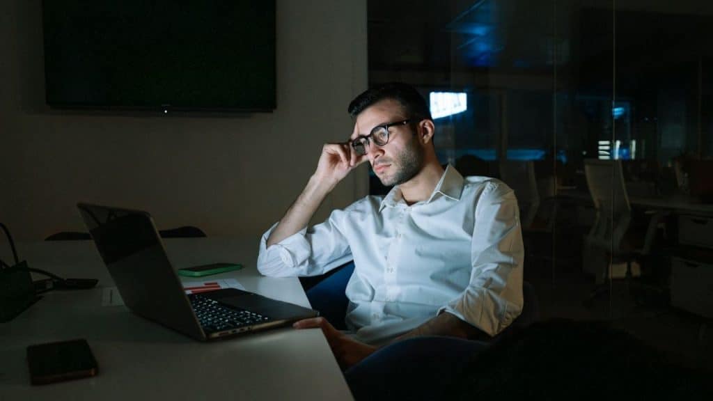 A man in glasses and a white shirt works alone on a laptop in a dark office.